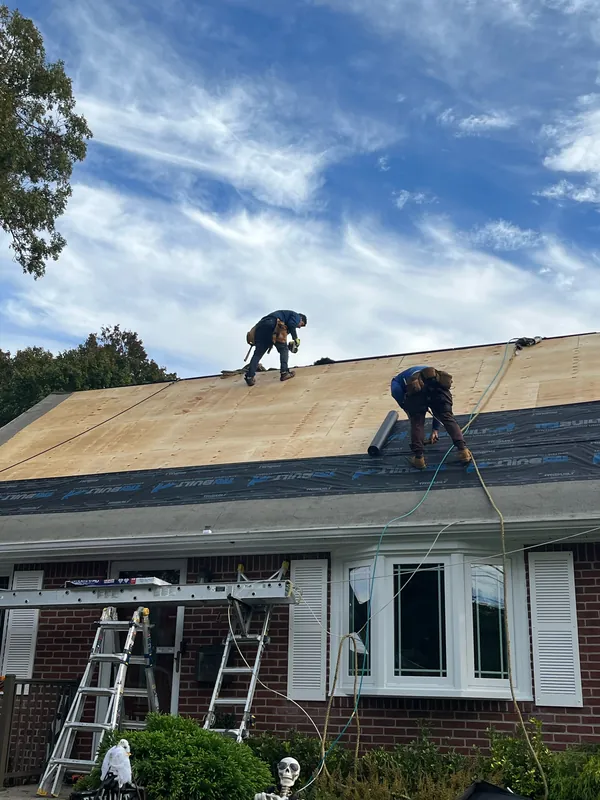 Two crew members on new plywood roof decking with black underlayment rolled out along the lower edge of a brick cape, ladders below.