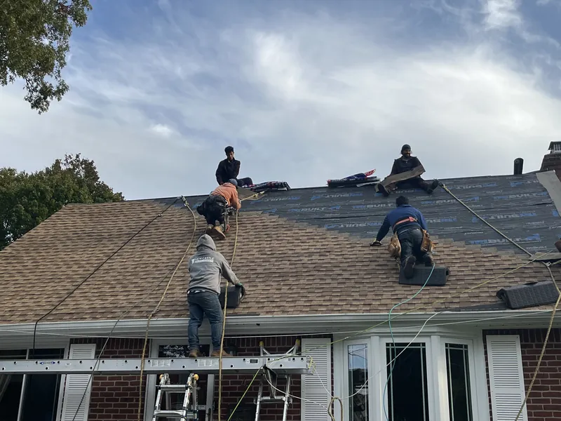 Crew installing tan shingles on a brick cape roof, with a worker on a ladder at the eave and underlayment still uncovered near the ridge.