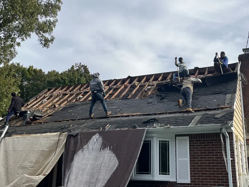Crew members on a brick cape roof tearing off the old shingles, with rafters exposed at the top and tarps hanging over the front.