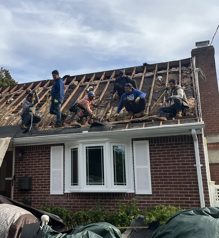 Crew members sitting and working on the bare rafters of a brick cape roof during tear-off, with a bay window and white gutter below.