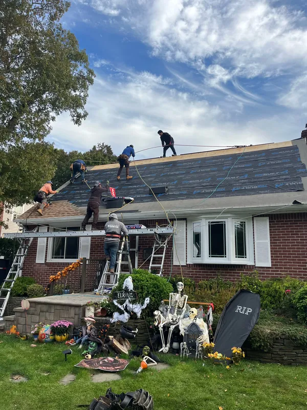 Crew climbing and working on a brick cape roof covered in black underlayment, with ladders and a ladder-jack plank at the eave and Halloween decorations on the lawn.