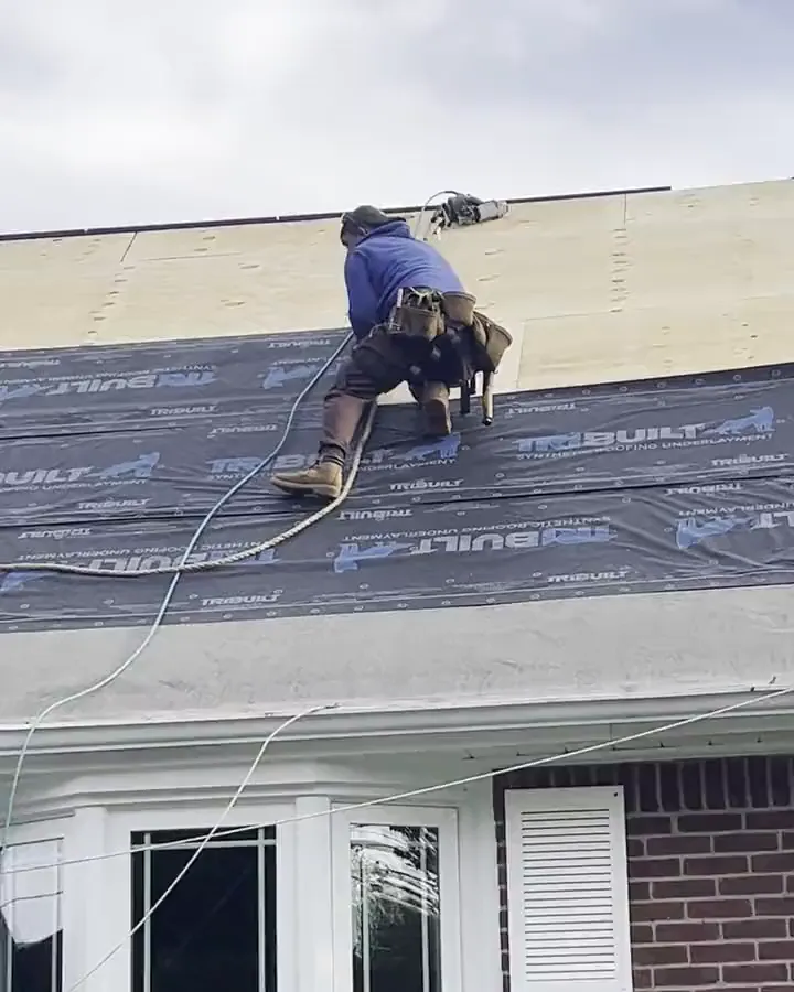 Crew working on a brick cape roof, laying underlayment below a strip of bare decking, with a worker at the top and a ladder-jack plank at the eave.