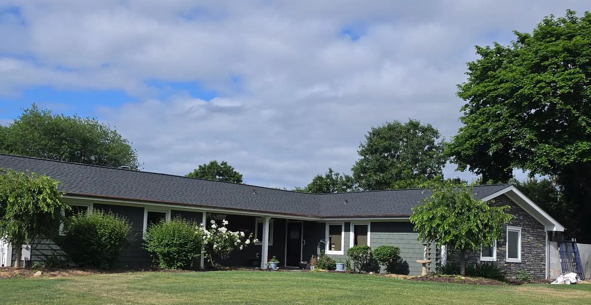 Wide view of the same ranch house with its new dark roof and a flowering shrub by the front.