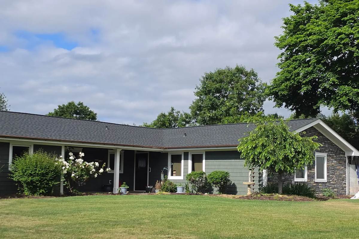 Gray ranch house with a new dark shingle roof, a green lawn in front and trees behind.
