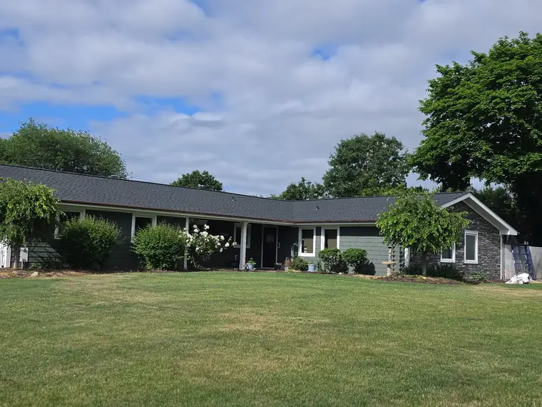 Wide view of the same ranch house with its new dark roof and a flowering shrub by the front.