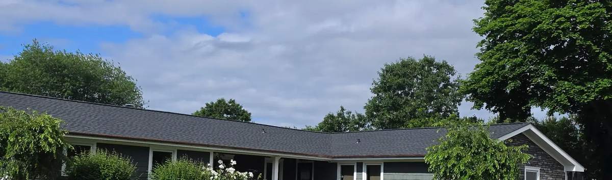 Wide view of the same ranch house with its new dark roof and a flowering shrub by the front.